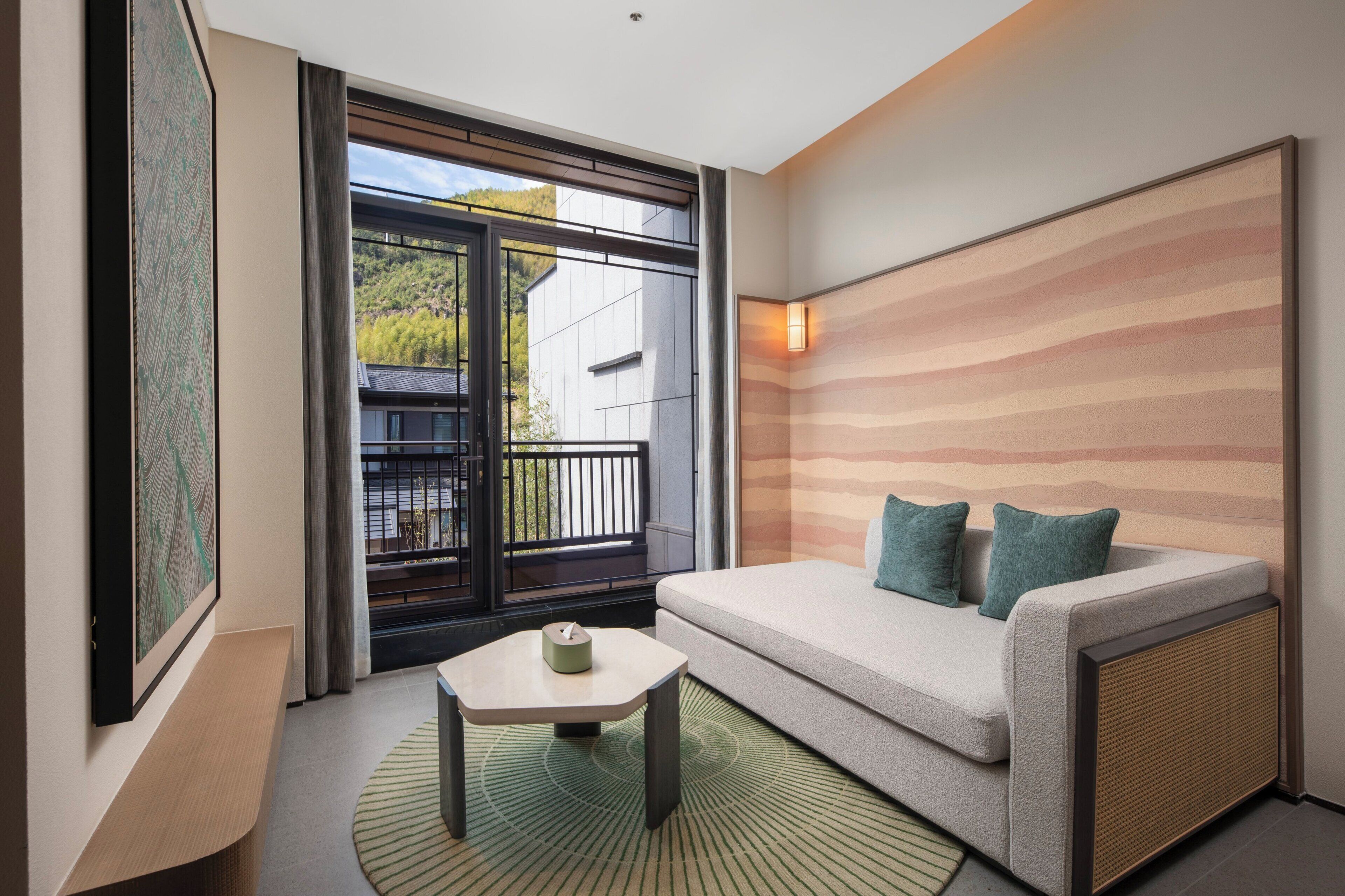 Ein modernes Hotelzimmer mit einer Couch, einem kleinen Tisch, einem Gemälde und Blick ins Freie.
