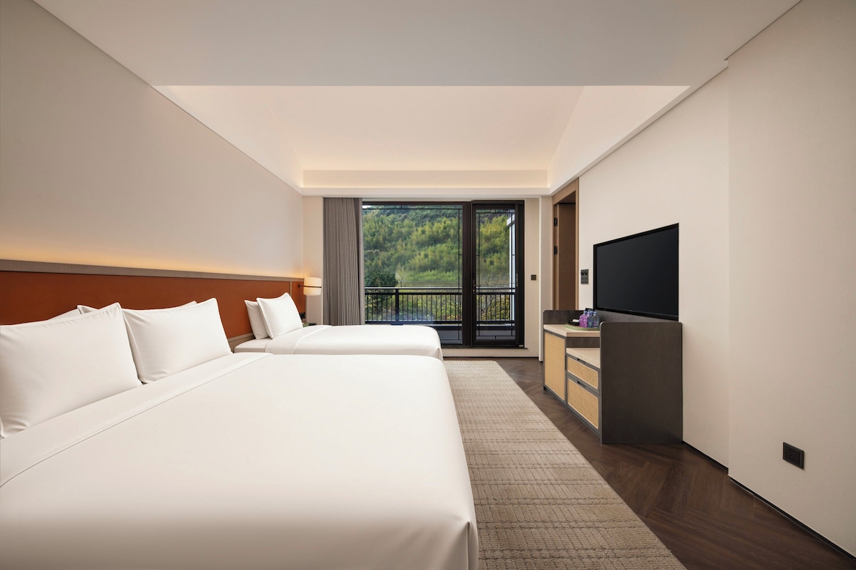 Une chambre d’hôtel avec deux lits, une télévision et un balcon donnant sur un paysage verdoyant.