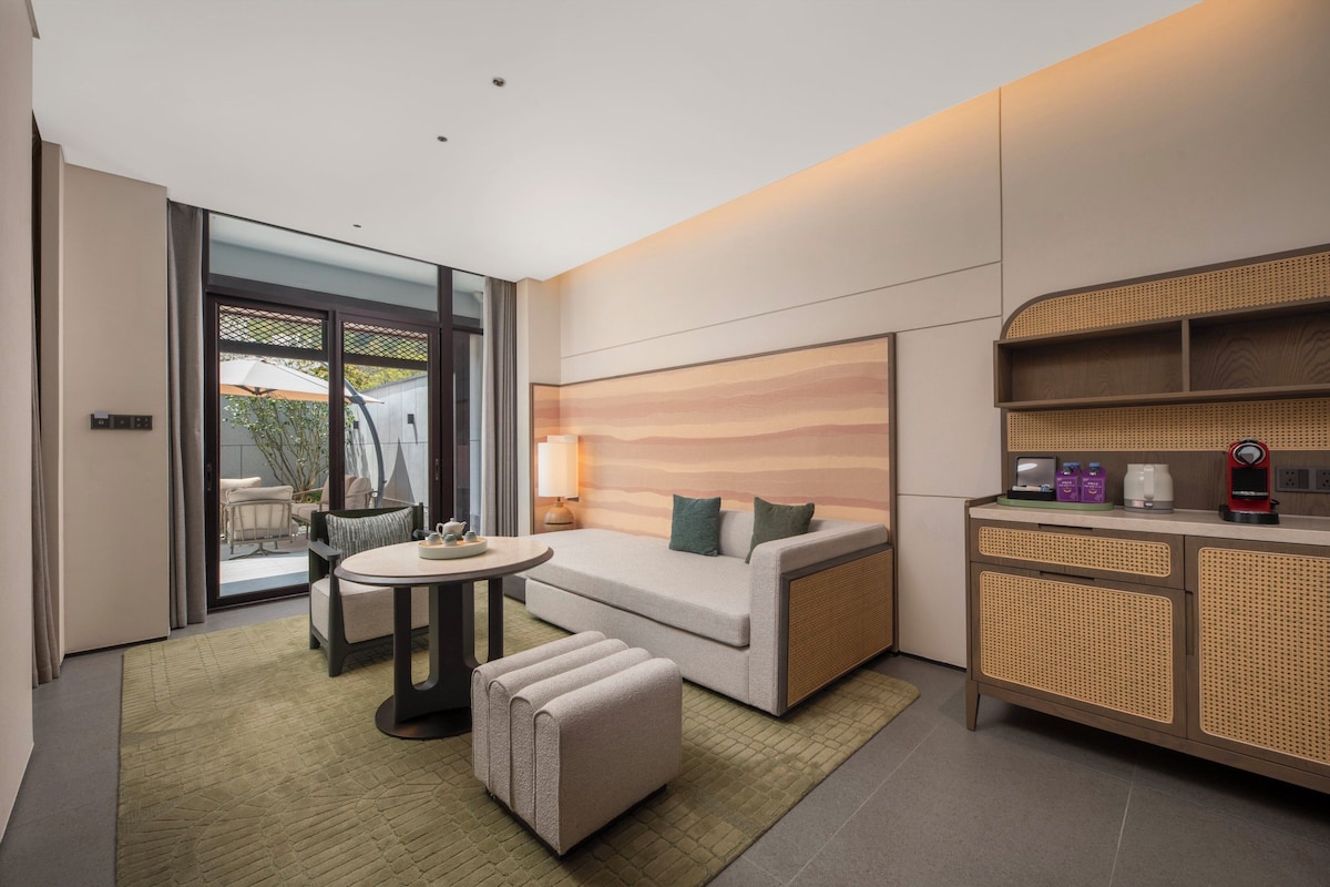 Une chambre d’hôtel moderne équipée d’un canapé, d’un pouf, d’une table à manger et d’un coin cuisine avec une machine à café.