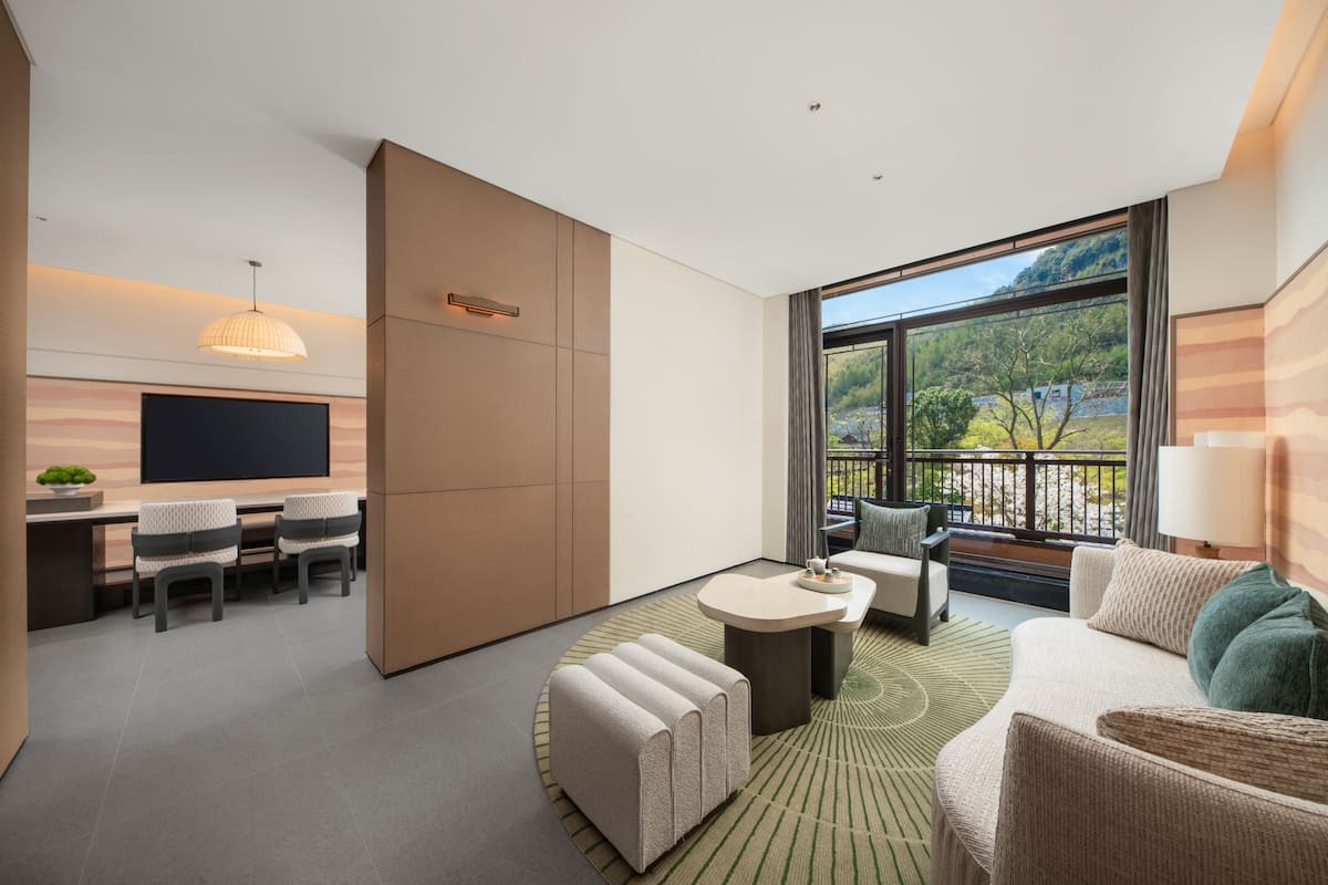 Una habitación de hotel moderna con televisión de pantalla plana, zona de comedor, sofá y ventana grande con vistas a zonas verdes.