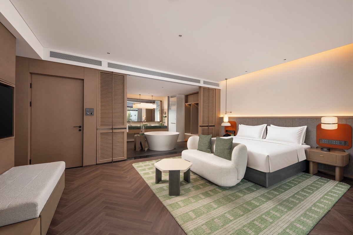 Une chambre d’hôtel moderne comprenant un lit, un canapé, une petite table, une salle de bain avec baignoire et un miroir.