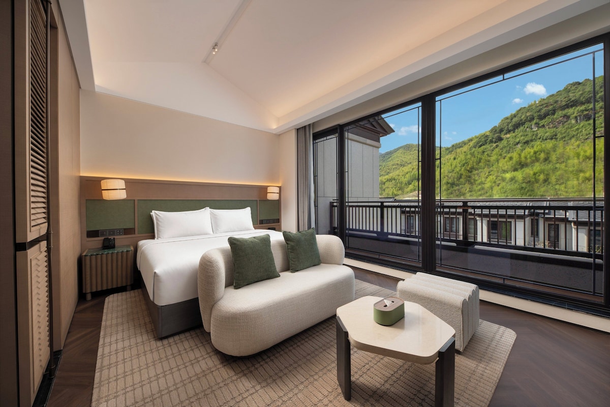 Une chambre d’hôtel moderne, dotée d’un grand lit, d’un canapé, d’une petite table et d’un balcon offrant une vue sur une colline verdoyante.