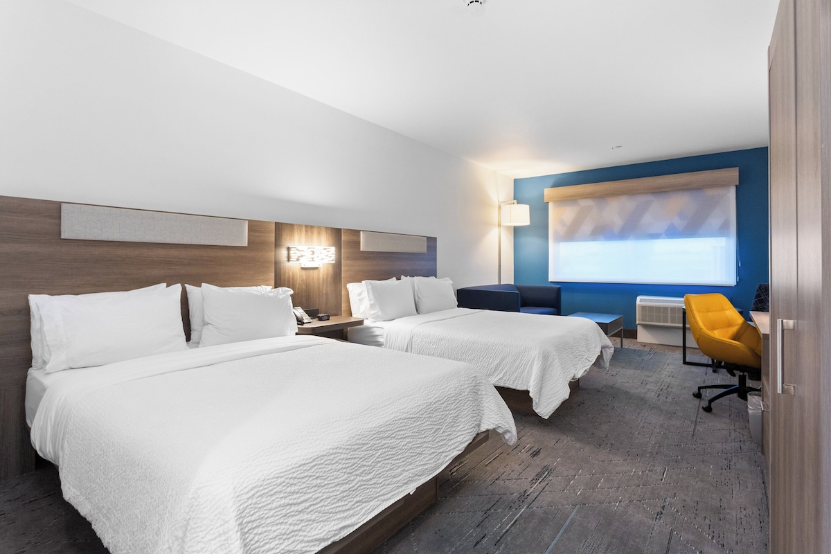 A hotel room with two beds, a desk, a chair, and a large window with blinds.