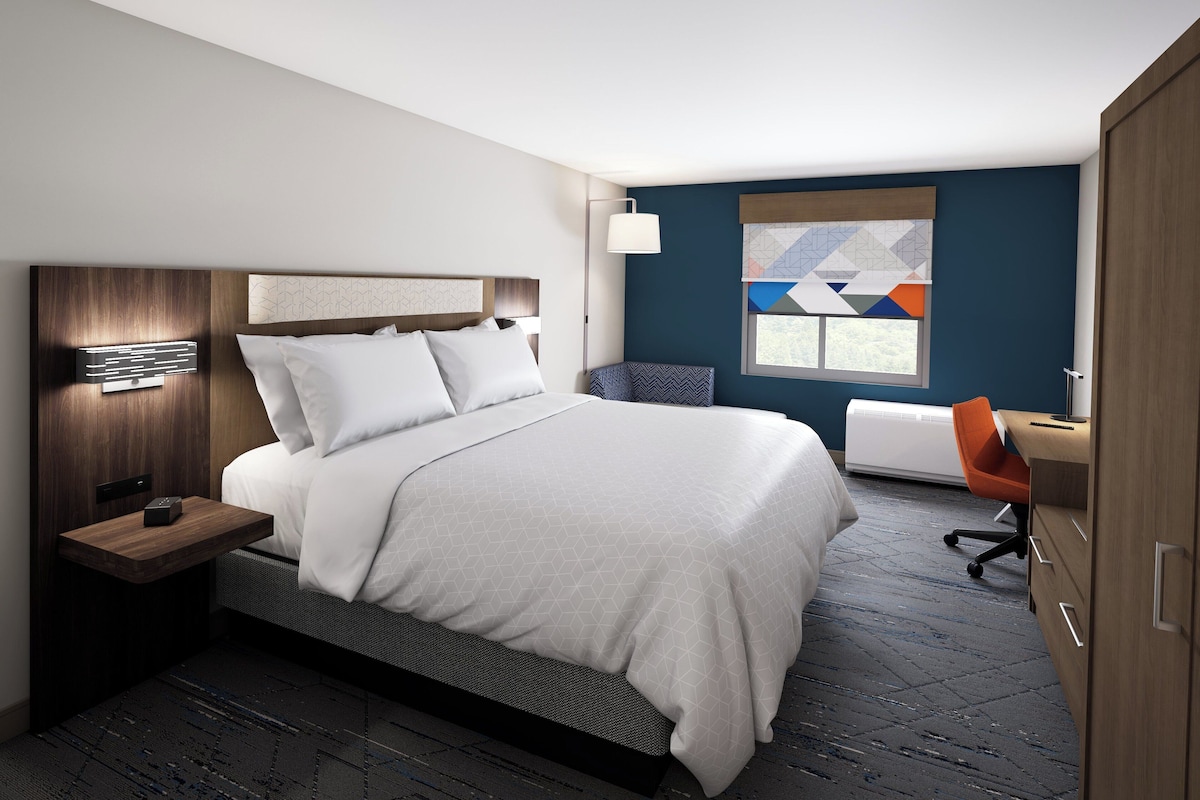 A modern hotel room with a large bed, a desk, and a window with a geometric view.