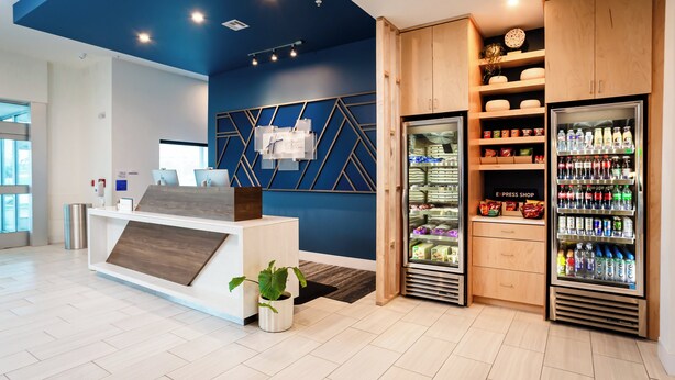A reception area with a reception desk, a wall-mounted computer, a refrigerator stocked with beverages, and a shelf with snacks. - Holiday Inn Express & Suites Waco Downtown by IHG (Waco)