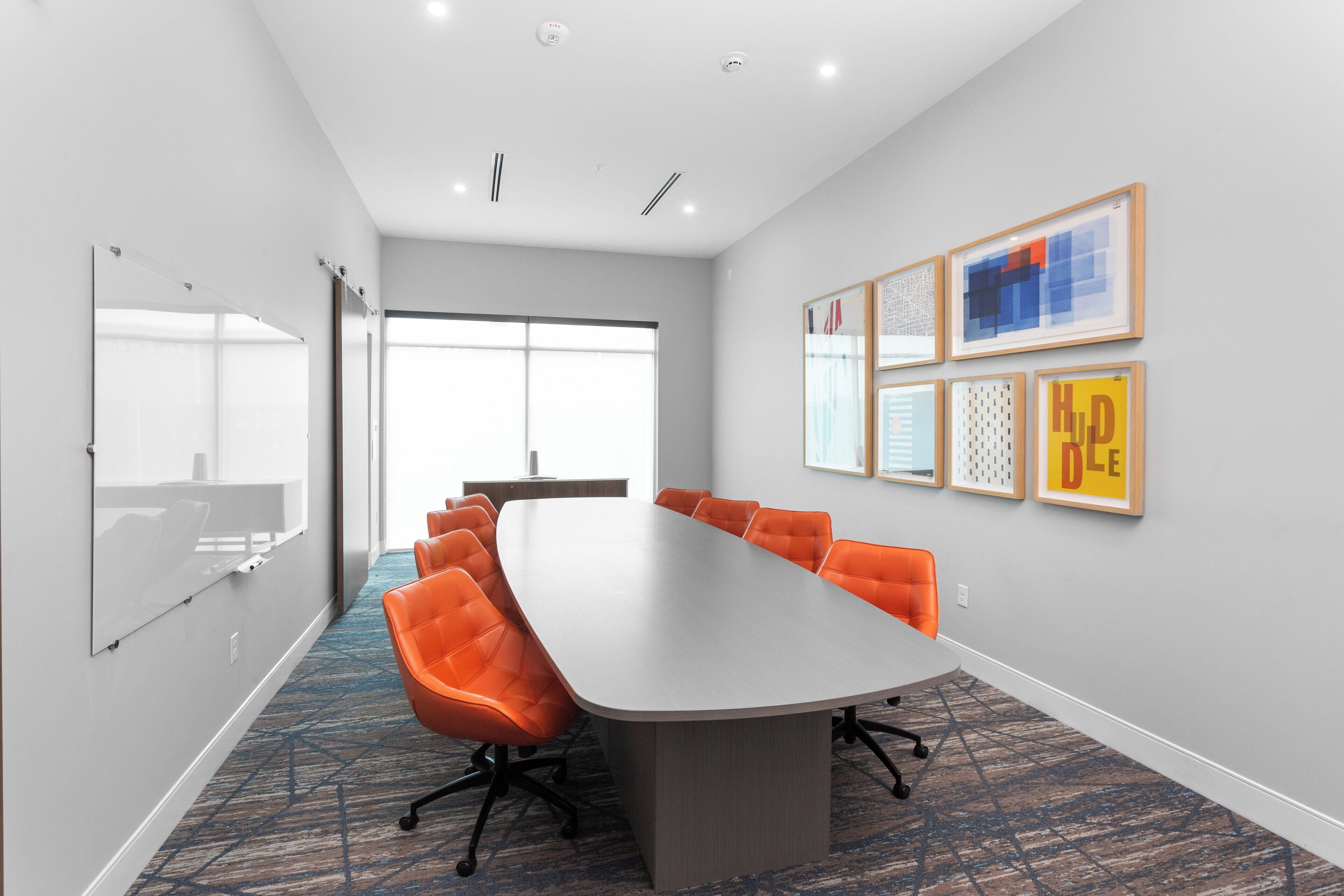 A conference room with a long table, orange chairs, a whiteboard, and framed artwork on the wall.