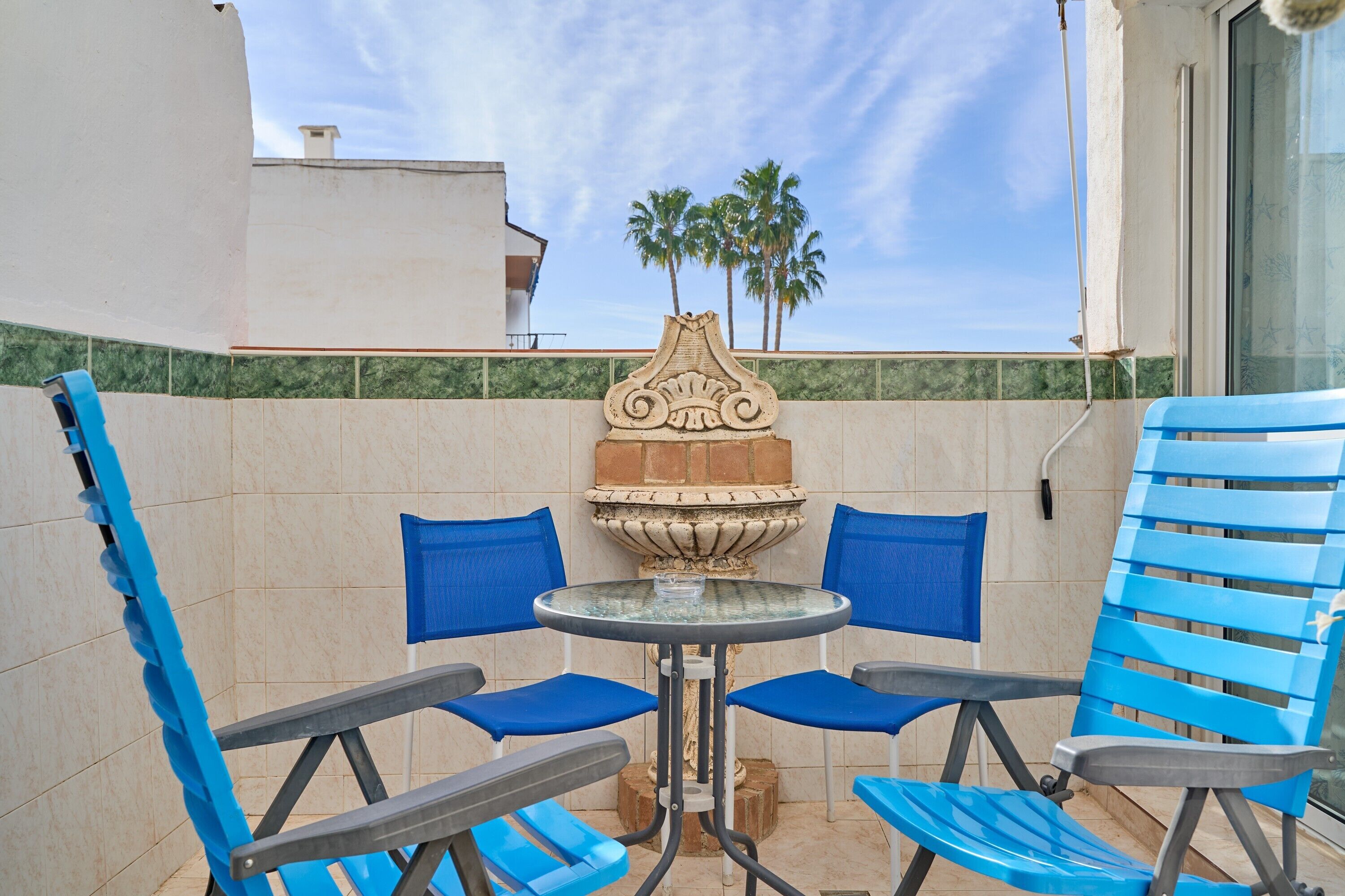 A patio with a round table and four blue chairs, a decorative stone fountain, and a view of palm trees.