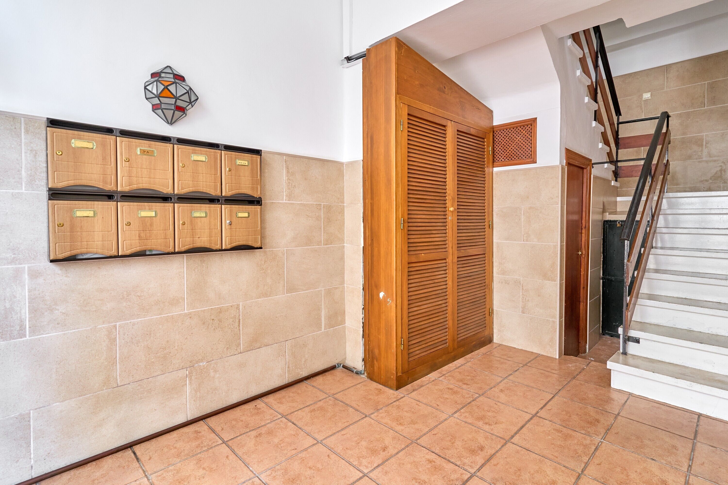 Un pasillo con suelo de baldosas, una puerta de madera, un armario empotrado en la pared y una escalera.