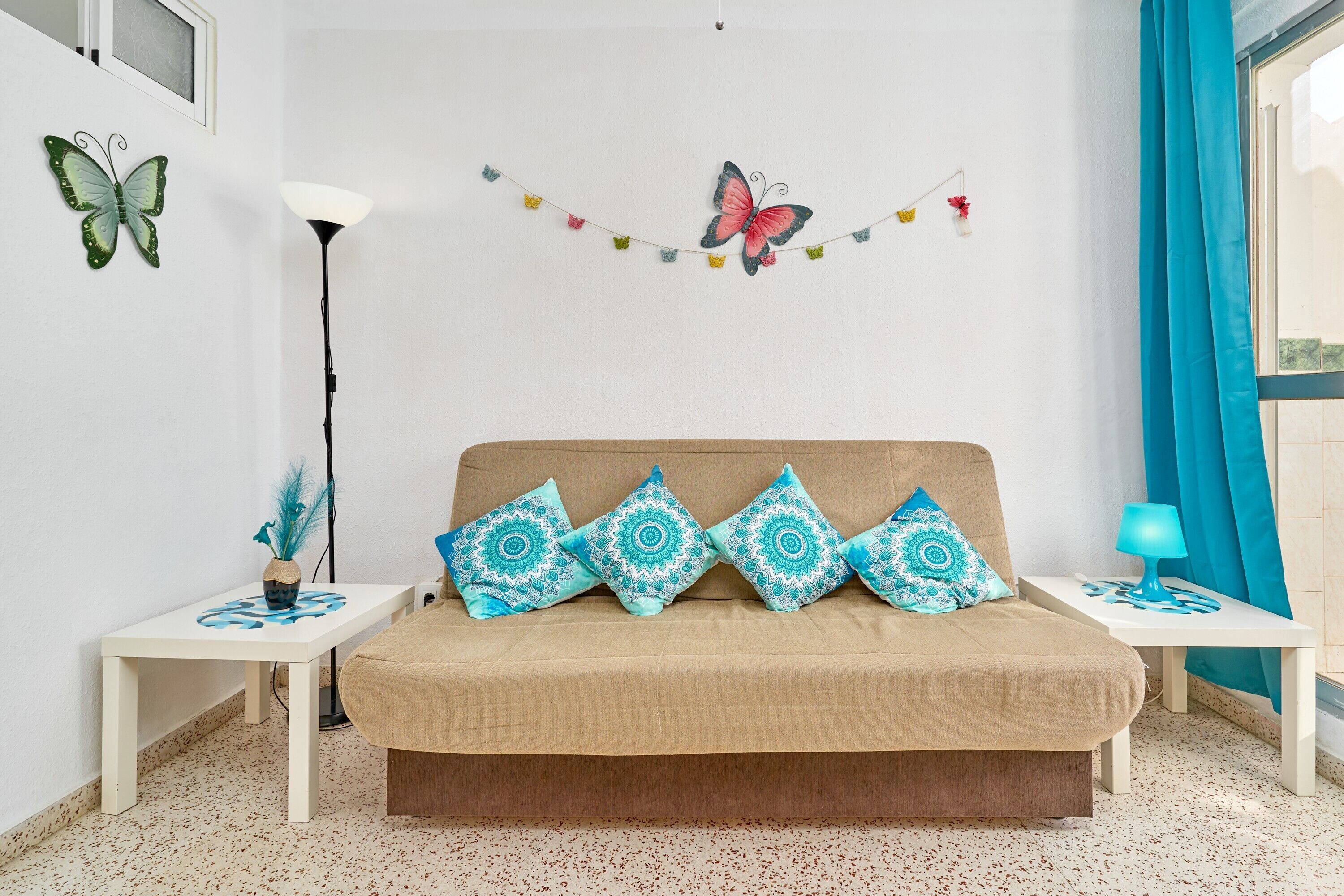 A room with a beige sofa, patterned pillows, a wall-mounted butterfly decoration, a string of colorful lights, a floor lamp, and a window with curtains.