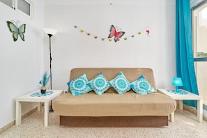 Een kamer met een beige bank, gekleurde kussens, een wanddecoratie van een vlinder, een lichtslingers, een vloerlamp en een raam met gordijnen.