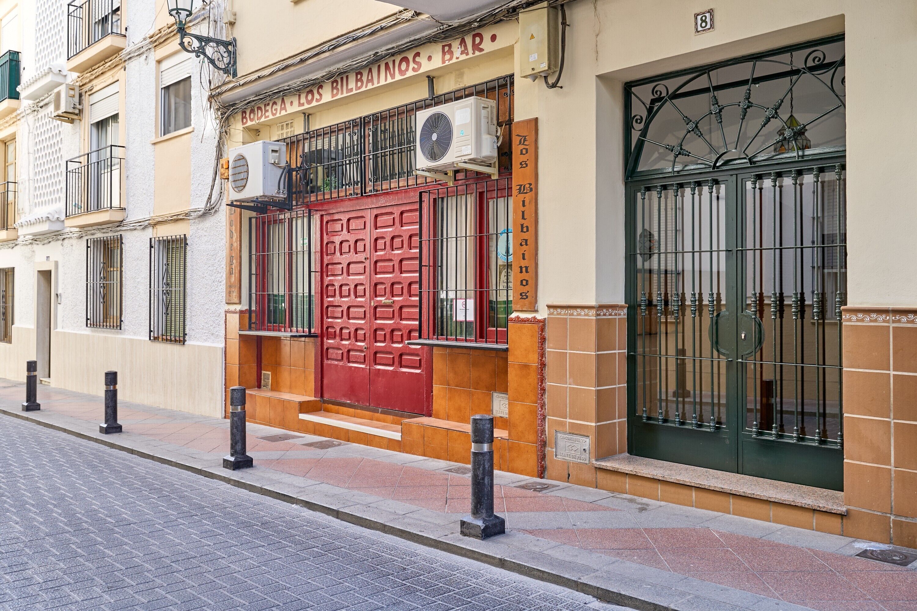Un edificio con puerta roja, unidades de aire acondicionado y una ventana con barrotes.