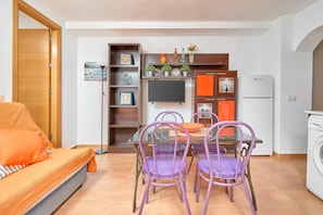 A compact living space with a sofa, dining table, purple chairs, a TV, shelves with decorative items, and a white washing machine.
