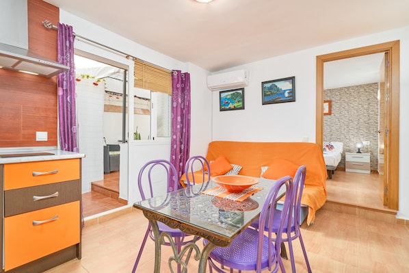 A compact room with a glass dining table, purple chairs, a bed with an orange cover, and a small kitchenette.