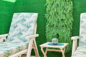 Two white chairs with palm leaf patterns, a small wooden table with a globe and a blue object, and a green wall with dense foliage.
