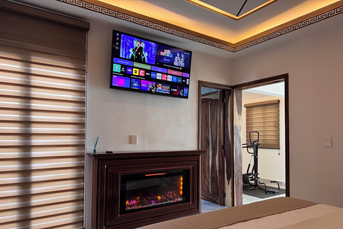 A hotel room with a flat-screen TV mounted above a fireplace, a bed with a beige cover, and a door leading to a balcony.