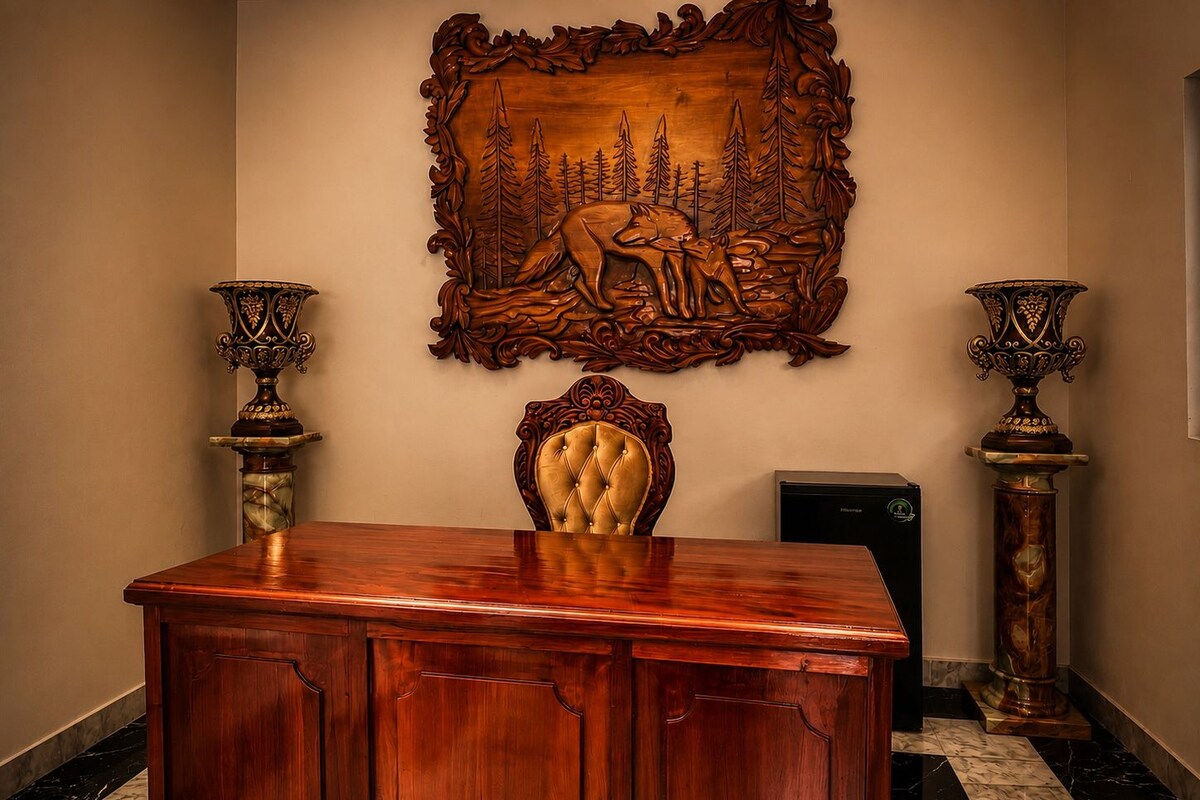 Un escritorio de madera con una silla tapizada, flanqueado por dos jarrones ornamentados, y una obra de arte de madera tallada arriba.