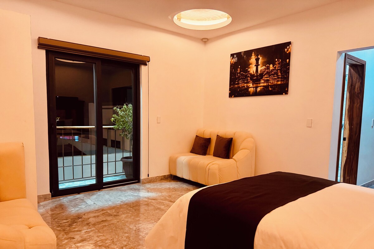 A hotel room with a bed, a sofa, a sliding glass door leading to a balcony, and a painting on the wall.