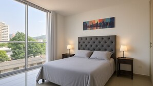 A modern bedroom with a large bed, two bedside tables with lamps, a cityscape painting on the wall, and a view of buildings through a large window.