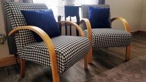 Two houndstooth patterned armchairs with wooden armrests and blue cushions, positioned in a room with a window and a small table with a plant.
