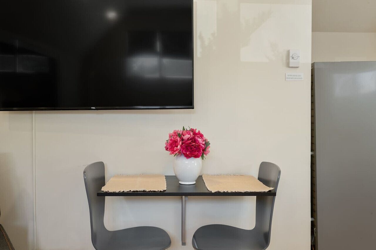 Una pequeña mesa con dos sillas, un jarrón blanco con flores rosadas, un televisor de pantalla plana montado en la pared y un refrigerador.