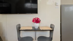 A small table with two chairs, a white vase with pink flowers, a flat-screen TV mounted on the wall, and a refrigerator.