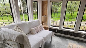 Una habitación acogedora con un sofá blanco, una lámpara y vistas a un jardín verde a través de amplios ventanales.