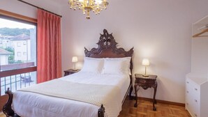 A bedroom with a large bed, two bedside tables with lamps, a chandelier, a window with curtains, and a view of buildings outside. - Holiday Home 'Vez' with Mountain View, Private Garden and Wi-Fi (Arcos de Valdevez)
