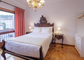 A bedroom with a large bed, two bedside tables with lamps, a chandelier, a window with curtains, and a view of buildings outside.