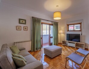 Un salon avec un canapé, un pouf, une télévision et une fenêtre avec des rideaux.