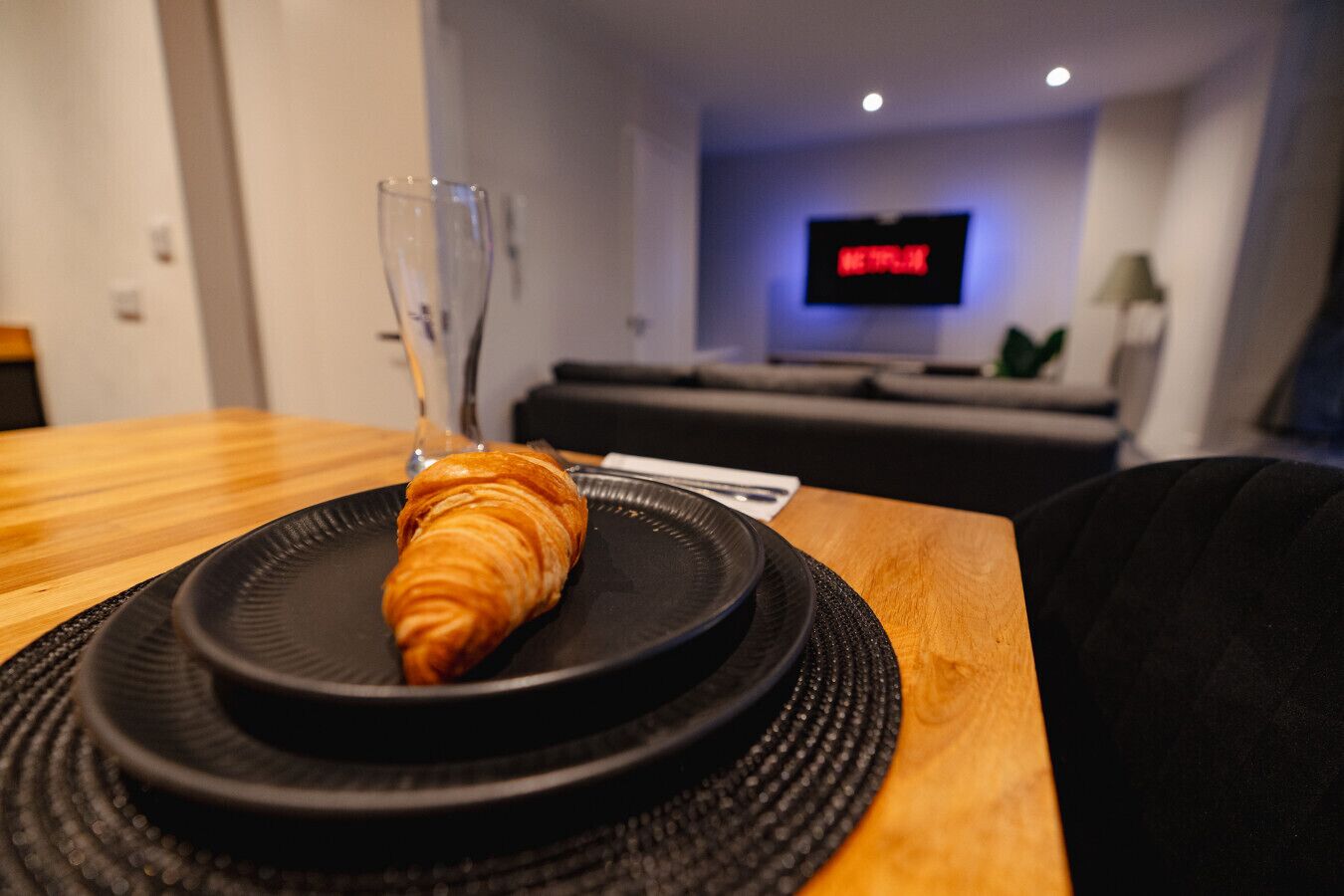 Ein Croissant auf einem schwarzen Teller auf einem Holztisch, im Hintergrund ein Fernseher mit Netflix-Anzeige.