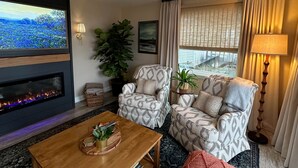 A living room with a fireplace, two patterned armchairs, a coffee table, and a large TV screen displaying a landscape. - Hutchins Lake Retreat (Fennville)