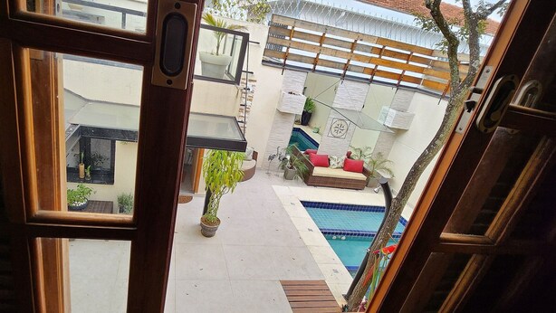 Vista a través de una ventana de madera que da a un patio con piscina, plantas en macetas y mobiliario exterior.