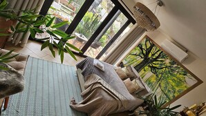 Una sala de estar acogedora con un sofá, un ventanal con vistas a la vegetación, una lámpara colgante y un cuadro de un árbol.