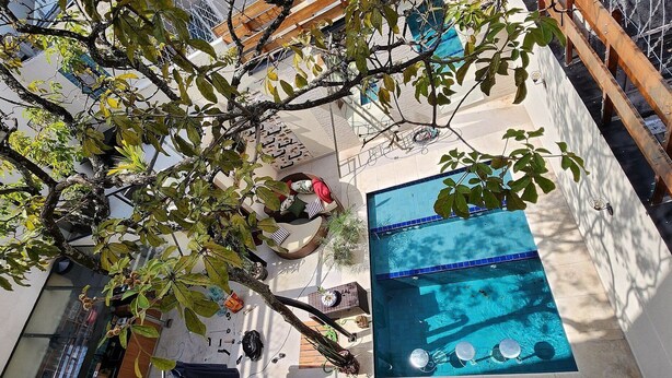 Un estrecho pasillo exterior con piscina, vigas de madera y un árbol.