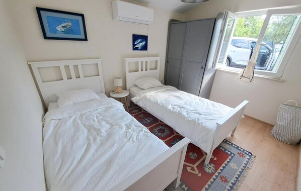 A room with two beds, a wardrobe, a window with a view of parked cars, and a small rug.