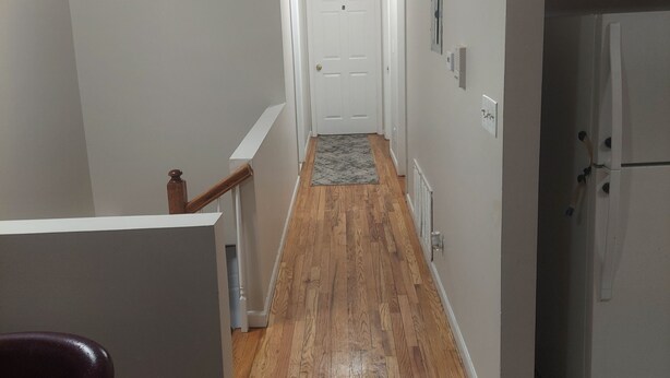 A narrow hallway with wooden flooring, white walls, and a white door at the end. - Crystal Room (Newark)
