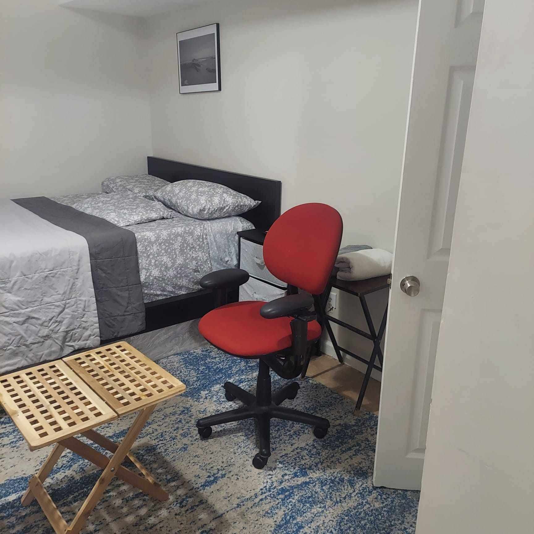 Un dormitorio con una cama, un escritorio, una silla de oficina roja y una pequeña mesa de madera.