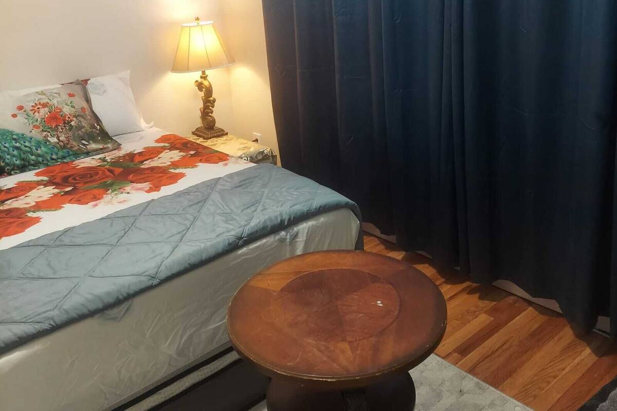 A bedroom with a bed, a nightstand, a lamp, a wooden stool, and a window with curtains.