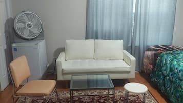 A small, furnished room with a white sofa, a single chair, a glass coffee table, a peacock-themed bedspread, a standing fan, and a small heater.