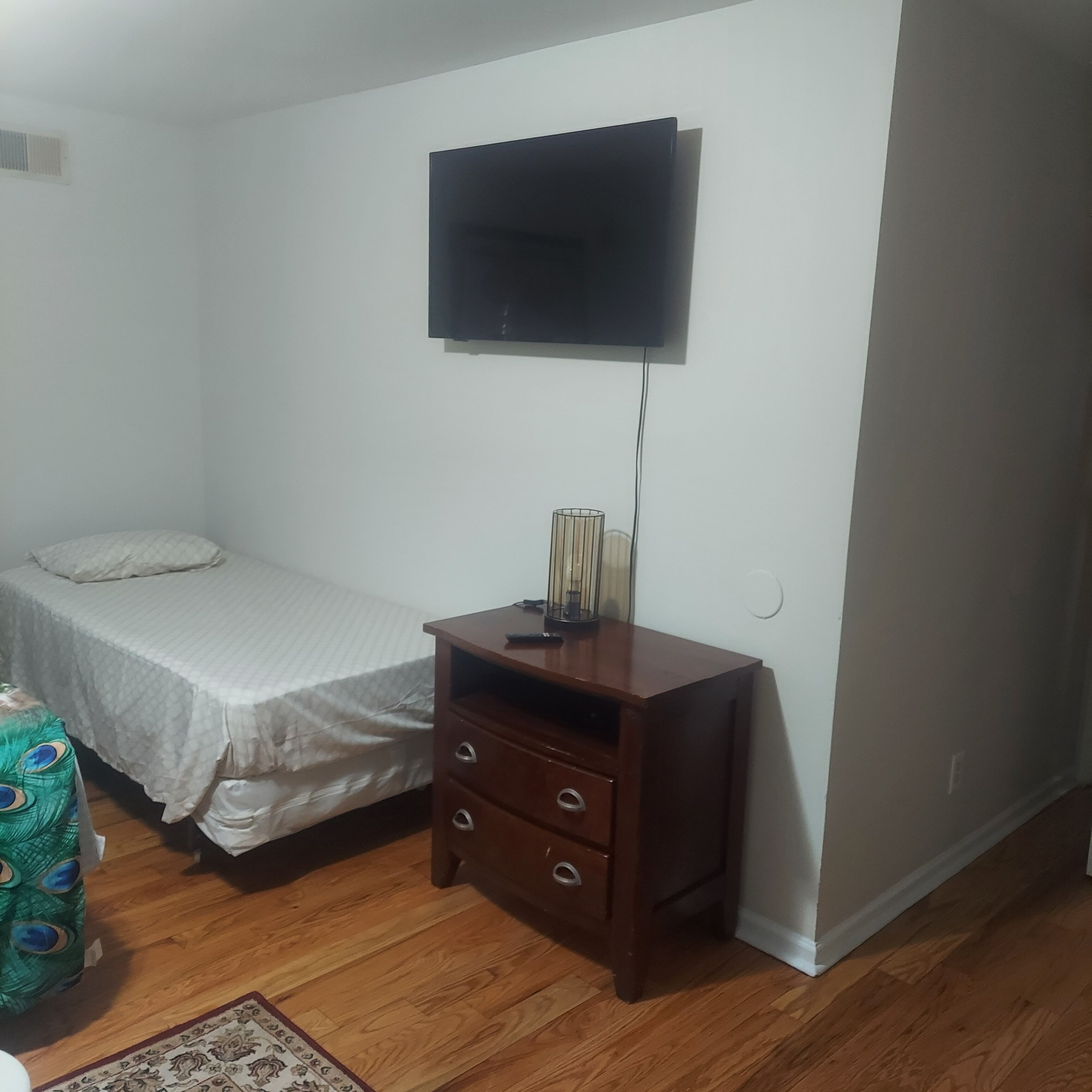 A small, sparsely furnished room with a bed, a wooden nightstand, a television mounted on the wall, and a patterned rug on the floor.