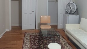 A room with a white sofa, a glass coffee table, a beige chair, and a standing fan. There are two white doors leading to closets. - Crystal Room (Newark)