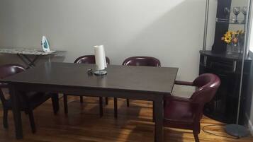 A dining area with a wooden table and four chairs, a small ironing board with an iron, a roll of paper towels, and a vase with flowers on a cabinet.