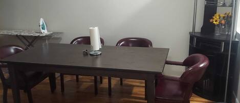 Een eetgedeelte met een houten tafel en vier stoelen, een klein strijkplank met strijkijzer, een rol keukenpapier en een vaas met bloemen op een kast.