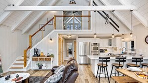 Un'ampia zona giorno in stile loft con soffitto alto, una scala in legno, una cucina con isola centrale e un'accogliente angolo salotto con divano in pelle.