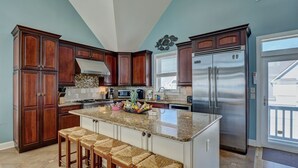 A kitchen with dark wood cabinets, a stainless steel refrigerator, a granite countertop, and a breakfast bar with woven stools. - Oceanfront Paradise with Amazing Views! (North Topsail Beach)