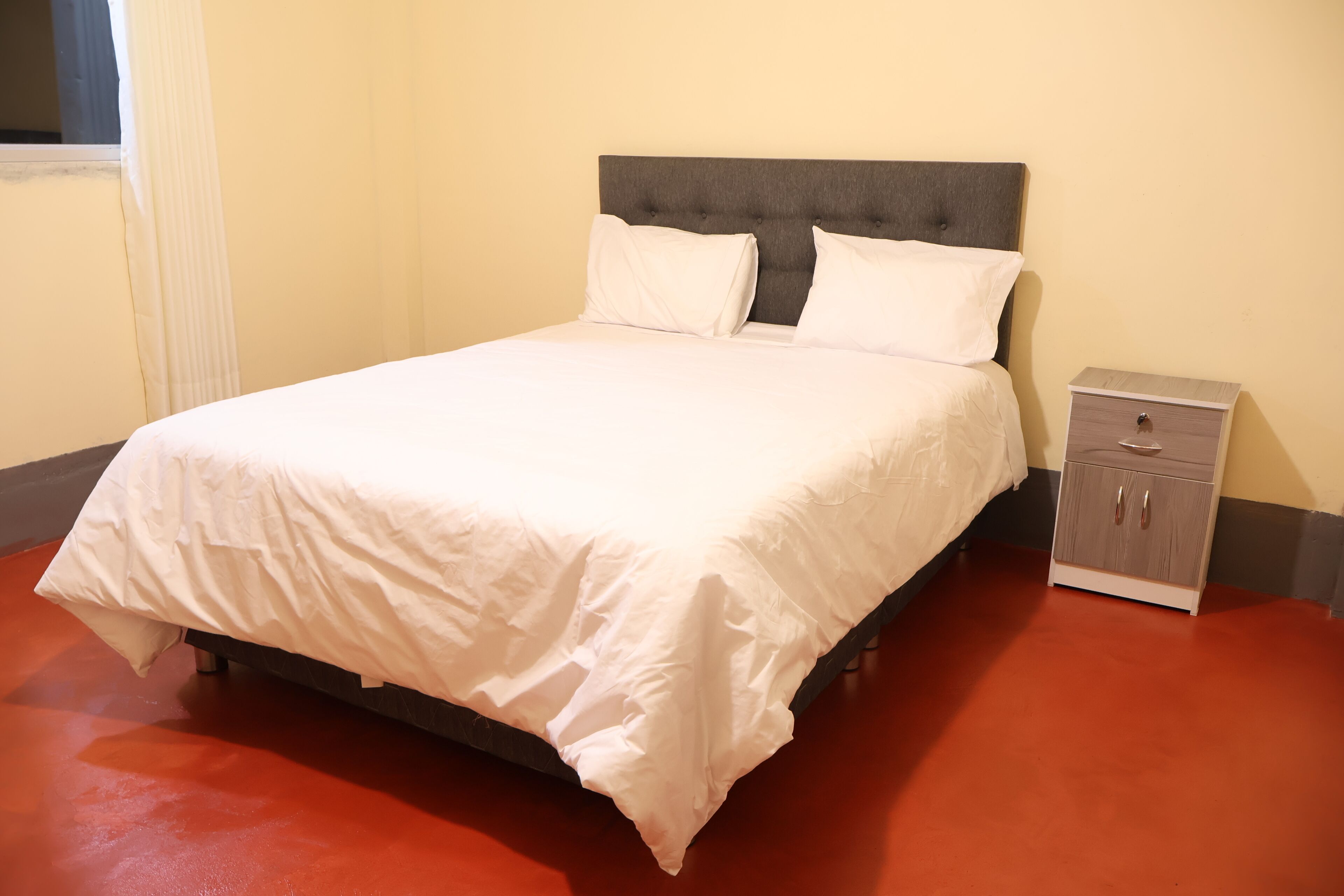 Cama bem arrumada com lençóis brancos, cabeceira estofada cinza e uma pequena mesa de cabeceira com gavetas.