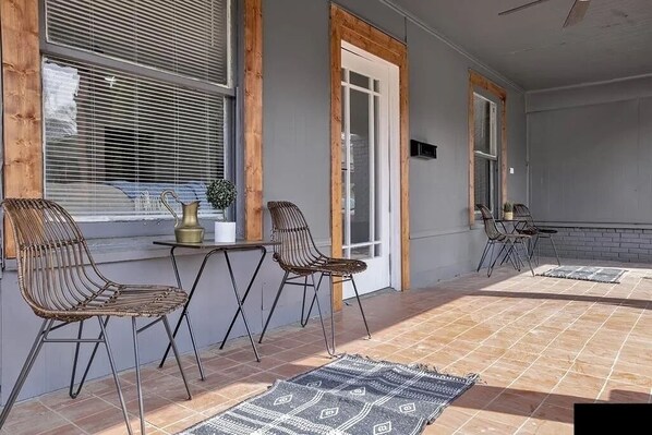 A sunlit patio with rattan chairs and a table, large windows with blinds, and a tiled floor.