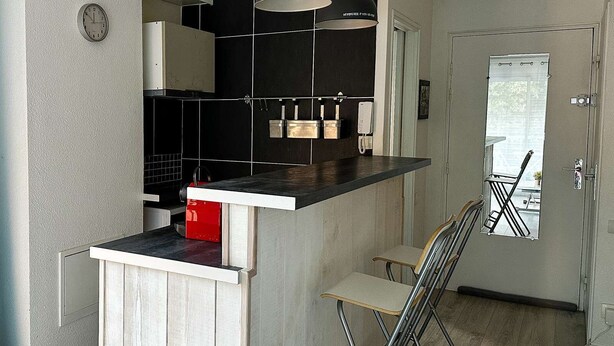 A compact kitchen with a black backsplash, a white countertop, and a wooden island. There are two bar stools and a folding chair. A clock and a phone are mounted on the wall. - Studio à Arcachon, emplacement idéal (ARCACHON)