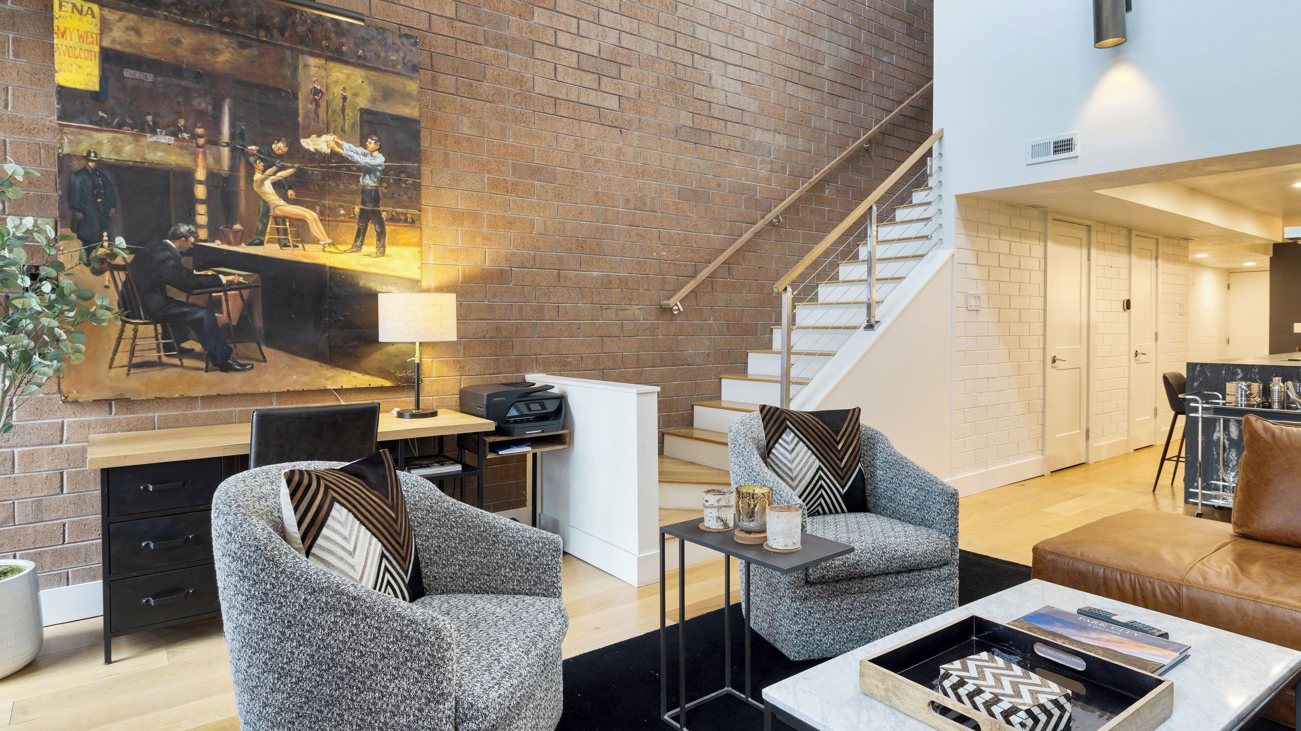 A modern living room with a brick wall, a painting, a staircase, and a kitchen area.