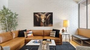A modern living room with a white brick wall, a brown leather sofa, a coffee table, a lamp, and a large ram artwork. - Parkside Condo by Avantstay Walk to Ski Lifts + Town Pool & Hot Tub Access (Park City)